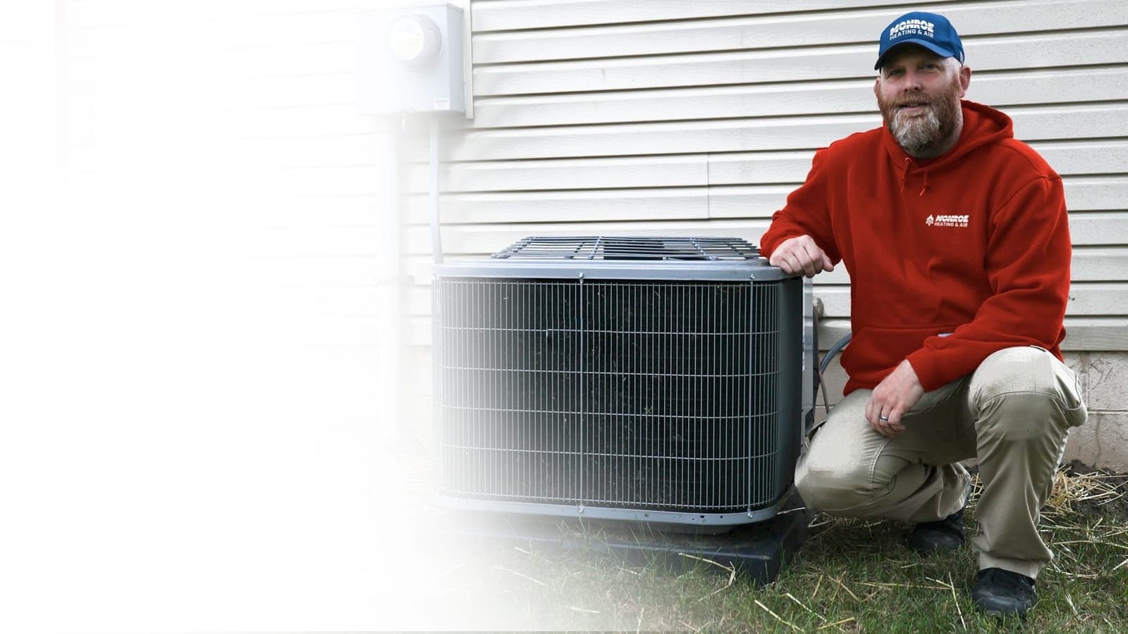 HVAC technician in a red hoodie and cap kneeling beside an outdoor air conditioning condenser unit next to a house with light siding